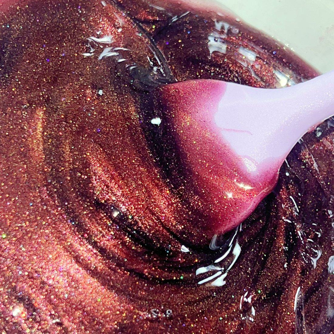 A lilac spatula stirring a glittering berry lip gloss mix in a glass bowl